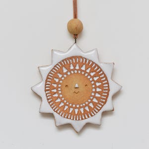 May include: A ceramic sun ornament with a white glazed border and a tan center featuring a stamped geometric pattern. The sun has a simple face with two dots for eyes and a small curved line for a mouth. It hangs from a brown cord with a wooden bead.