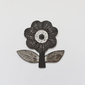 Op de afbeelding: Een plat, decoratief bloemornament met een zwart, getextureerd oppervlak met witte geëtste lijnen. Het midden van de bloem is een witte cirkel met een cluster van kleine zwarte stippen. Twee symmetrische bladeren steken uit de centrale steel.