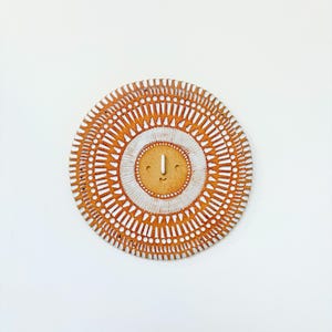 May include: Circular ceramic wall art with a sunburst design. The piece has a light brown base with white accents and a central face-like design. The diameter is approximately 25 cm.