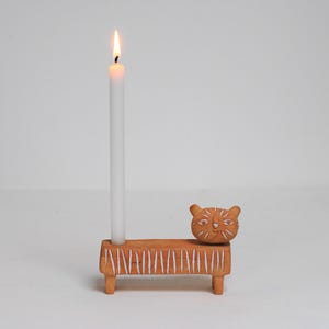 Puede incluir: Un candelabro de madera con forma de gato con una vela blanca encendida. El soporte es de color marrón claro con rayas verticales blancas y una cabeza de gato tallada. La vela es alta y delgada, con una pequeña llama en la parte superior.
