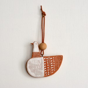 May include: A ceramic bird ornament featuring a white glazed head and chest with a terracotta-colored body decorated with white geometric patterns. The piece hangs from a brown leather cord accented with a single round wooden bead at the top.