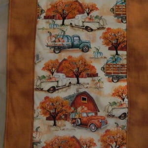 Puede incluir: Un camino de mesa de tela rectangular con un panel central de estampado otoñal repetitivo de camiones antiguos, calabazas y graneros en tonos naranja, marrón y azul verdoso, bordeado por tiras de tela marrón moteada a ambos lados.