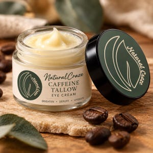 Puede incluir: Un frasco de vidrio de crema para ojos Natural Craze Caffeine Tallow con una crema de color amarillo pálido en su interior. La etiqueta muestra el texto Natural Craze, Caffeine Tallow Eye Cream, Brighten, Depuff, Hydrate. Una tapa verde oscuro con un logo de hoja está al lado.
