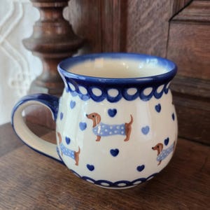 Może przedstawiać: Ceramiczny kubek o zaokrąglonym kształcie z wzorem brązowych jamników w jasnoniebieskie sweterki w kropki, przeplatany małymi niebieskimi sercami. Krawędź i uchwyt są ciemnoniebieskie, a podstawa ma niebieski, falisty wzór.
