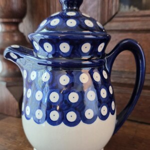 May include: A ceramic teapot with a lid, featuring a dark blue and white polka dot pattern. The teapot has a rounded body, a curved handle, and a short spout. The upper portion of the body and the lid are decorated with dark blue circles containing white centers, while the lower portion of the teapot remains a solid, plain cream-white color.
