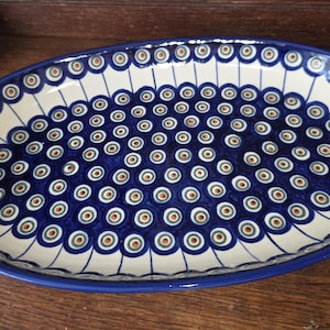 May include: An oval ceramic serving platter featuring a repeating peacock eye pattern in shades of deep blue, green, and brown on a cream-colored background, with a solid blue rim and radiating line details along the outer edge.