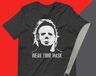 May include: A dark grey t-shirt featuring a white silhouette of Michael Myers from the Halloween films wearing his iconic mask. The text "WEAR YOUR MASK" is printed below the image.