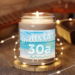 Peut inclure: Une bougie en verre brun avec une étiquette blanche qui dit "Smells like 30a". L'étiquette représente une scène de plage bleue et blanche avec un soleil jaune.