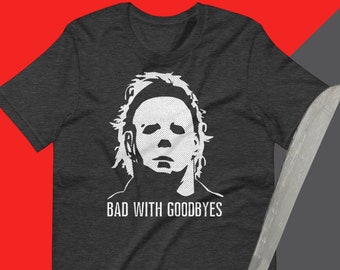 May include: A dark grey t-shirt featuring a white graphic of Michael Myers from the Halloween films. The graphic includes the text "Bad with Goodbyes".