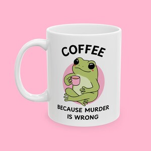 Puede incluir: Taza de cerámica blanca con una ilustración de una rana verde sosteniendo una taza de café. La taza tiene texto negro que dice "Coffee because murder is wrong."