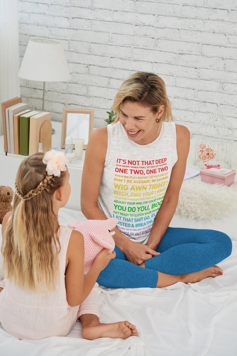K&ouml;nnte beinhalten: Eine Frau tr&auml;gt ein graues Tanktop mit einem bunten Text-Motiv, auf dem "It's not that deep... One Two. One Two... Wig Awn Tight!" steht. Sie sitzt auf einem Bett mit einem jungen M&auml;dchen. Die Frau l&auml;chelt und schaut das M&auml;dchen an. Das M&auml;dchen h&auml;lt ein rosa geh&auml;keltes Spielzeug in der Hand.