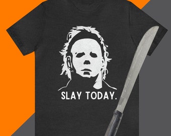Może przedstawiać: Szara koszulka z nadrukiem białej grafiki maski Michaela Myersa i napisem "SLAY TODAY". Na koszulce leży czarny maczeta.