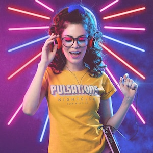 May include: A woman with curly brown hair is wearing a yellow t-shirt with the text "Pulsations Nightclub Glen Mills, PA". She is wearing headphones and singing along to music. The background is a purple and blue gradient with neon pink and blue lights.