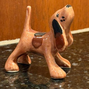 May include: A brown ceramic dog figurine with black and brown accents. The dog is standing on all fours with its head tilted upwards. The figurine has a glossy finish and is likely a decorative item.