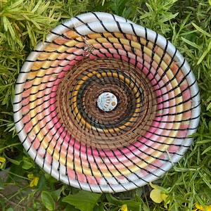 May include: A woven basket with a circular shape and a colorful design. The basket is made of natural materials and has a silver trim. The basket is surrounded by green plants.