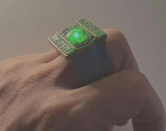 May include: A metallic ring featuring a dark, textured band and a rectangular top section. The center of the ring contains a circular, glowing green light element surrounded by a detailed, raised border with a metallic finish.