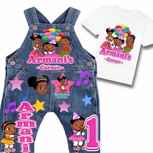 Op de afbeelding: Blauwe spijker tuinbroek en een wit t-shirt met kleurrijke cartoonafbeeldingen van kinderen, ballonnen en sterren. De tekst luidt Armani's Corner en Gracie's Corner, met een groot roze cijfer één op de broekspijp van de tuinbroek.