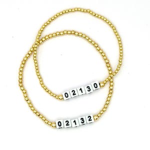 May include: Two gold-toned beaded bracelets featuring small, round metallic beads. Each bracelet includes a segment of white square beads with black printed numbers. One bracelet displays the sequence 02130, and the other displays 02132.