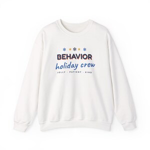May include: A white long-sleeve sweatshirt featuring the text BEHAVIOR in dark blue with an orange outline, holiday crew in blue script, and JOLLY - PATIENT - KIND in black, accented by blue and yellow snowflake and star icons.