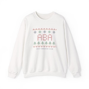 May include: A white crewneck sweatshirt featuring a festive knit-style graphic print. The design includes red, green, and blue patterns of small trees and geometric shapes, the letters ABA in red, and the text UGLY SWEATER CLUB in black.