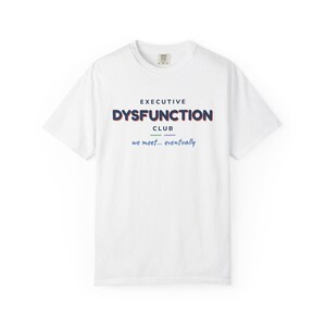 May include: A white short-sleeve t-shirt featuring the text "EXECUTIVE DYSFUNCTION CLUB" in dark blue with a red shadow effect. Below the text are two small horizontal lines in green and purple, followed by the phrase "we meet... eventually" in blue.