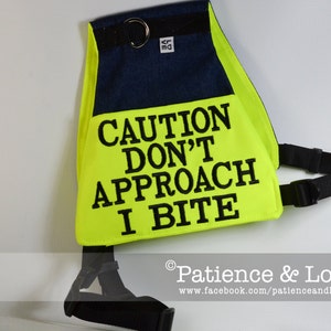 Könnte beinhalten: Eine leuchtend gelbe Hundeweste mit schwarzem Text, der "CAUTION DON'T APPROACH I BITE" lautet. Die Weste hat einen schwarzen Gurt und ein blaues Jeans-Patch mit einem silbernen Ring.