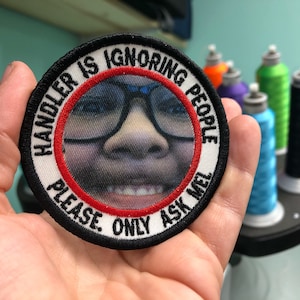 Könnte beinhalten: Runder, gestickter Aufnäher mit dem Text "HANDLER IS IGNORING PEOPLE. PLEASE. ONLY ASK MEL." Der Aufnäher zeigt eine Person mit Brille. Der Aufnäher hat einen schwarzen Rand mit einem roten Kreis.