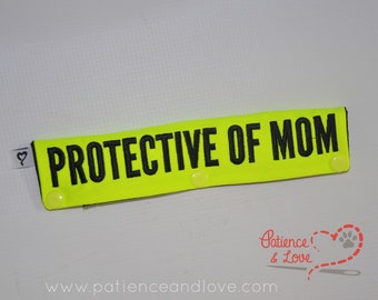 Peut inclure: Un patch en tissu jaune vif avec du texte noir qui dit "PROTECTIVE OF MOM". Le patch a plusieurs petits boutons-pression en plastique transparent.