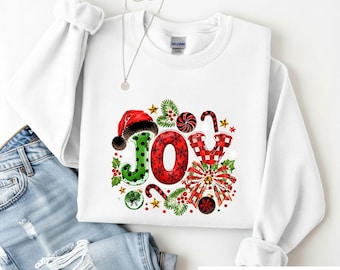 May include: A white long-sleeve sweatshirt featuring a graphic print of the word JOY. The letter J is green with black polka dots and a Santa hat, the O is red with black snowflakes, and the Y is red and white buffalo plaid.