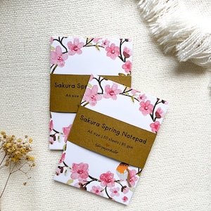 Puede incluir: Dos blocs de notas Sakura Spring de tamaño A6 con 50 hojas de papel de 80 g/m². Los blocs de notas presentan un diseño de acuarela con flores de cerezo rosas y un petirrojo en una rama. Los blocs de notas están envueltos en papel marrón con el texto "Sakura Spring Notepad" y "twinpaperstudio" impreso en ellos.