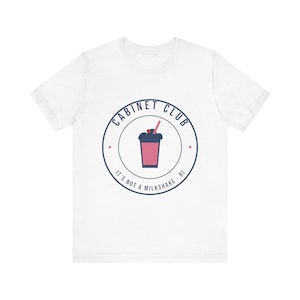 Może przedstawiać: Biały t-shirt z krótkim rękawem z okrągłym logo na piersi. Logo zawiera różowy napój ze słomką i wisienką, otoczony napisem CABINET CLUB oraz IT'S NOT A MILKSHAKE · RI wykonanym ciemnoniebieskimi literami.