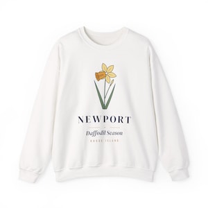 May include: A white crewneck sweatshirt featuring a graphic of a yellow daffodil with green leaves. Below the flower, the text reads NEWPORT, Daffodil Season, and RHODE ISLAND in dark blue and tan lettering on the front of the garment.