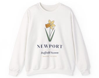 May include: A white crewneck sweatshirt featuring a graphic of a yellow daffodil with green leaves. Below the flower, the text reads NEWPORT, Daffodil Season, and RHODE ISLAND in dark blue and tan lettering on the front of the garment.