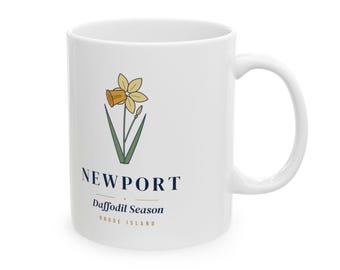May include: A white ceramic mug featuring a yellow daffodil illustration with green stems. Below the flower, the text reads NEWPORT, Daffodil Season, and RHODE ISLAND in dark blue and tan lettering, centered on the front of the mug.