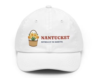 Peut inclure: Une casquette de baseball blanche ornée d'un motif représentant un panier tressé marron rempli de jonquilles jaunes. À droite du panier, le texte NANTUCKET est imprimé en rouge, avec les mots DAFFODILS BY THE BASKETFUL en noir en dessous.