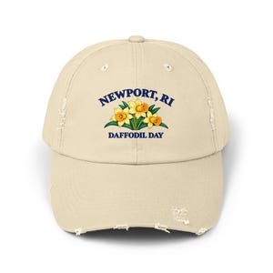 May include: A beige distressed baseball cap featuring a graphic of three yellow daffodils with green leaves. The text "NEWPORT, RI" is printed in dark blue above the flowers, and "DAFFODIL DAY" is printed in dark blue below the flowers.