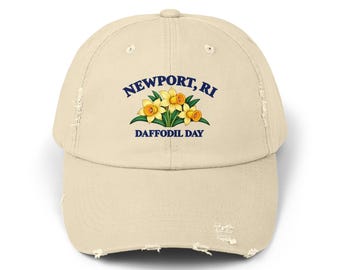 May include: A beige distressed baseball cap featuring a graphic of three yellow daffodils with green leaves. The text "NEWPORT, RI" is printed in dark blue above the flowers, and "DAFFODIL DAY" is printed in dark blue below the flowers.