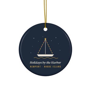 May include: A round dark navy blue ceramic ornament with a gold hanging string. The center features a white sailboat graphic with a star and string lights on the mast. Text reads: Holidays by the Harbor, NEWPORT · RHODE ISLAND.