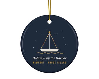 May include: A round dark navy blue ceramic ornament with a gold hanging string. The centre features a white sailboat graphic with a star and string lights on the mast. Text reads: Holidays by the Harbor, NEWPORT · RHODE ISLAND.