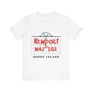 May include: A white short-sleeve t-shirt featuring a graphic of a cartoon character peeking over a brick wall. The red text reads NEWPORT WAZ 'ERE, with the words RHODE ISLAND printed in black block letters underneath the brick wall design.
