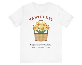 Puede incluir: Una camiseta blanca de manga corta con un gráfico de una cesta tejida llena de narcisos amarillos. El texto NANTUCKET está arqueado encima, con Daffodils by the Basketful y ACK ISLAND SPRING impresos debajo de la cesta.