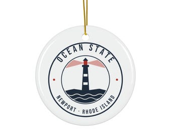 May include: A round white ceramic ornament with a gold hanging string. The design features a dark blue lighthouse graphic with red light beams, set above stylized waves. Text reads OCEAN STATE and NEWPORT RHODE ISLAND in dark blue.