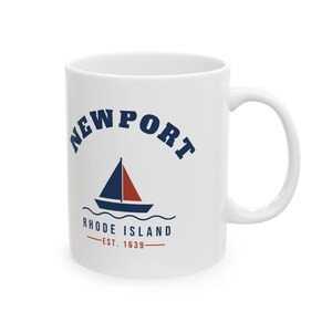 May include: A white ceramic mug featuring a navy blue and red graphic of a sailboat on waves. The text reads NEWPORT in an arch above the boat, with RHODE ISLAND and EST. 1639 printed in navy blue and red below the sailboat design.