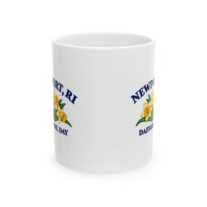 May include: A white ceramic mug featuring a graphic of yellow daffodils with green leaves. The text in dark blue reads Newport, RI and Daffodil Day. The design is printed on the side of the cylindrical mug with a smooth, glossy finish.