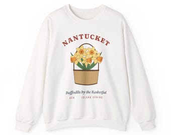 Puede incluir: Una sudadera blanca de cuello redondo con un gráfico de narcisos amarillos en una cesta tejida marrón. El texto dice NANTUCKET en rojo, seguido de Daffodils by the Basketful y ACK - ISLAND SPRING en letras negras y rojas.