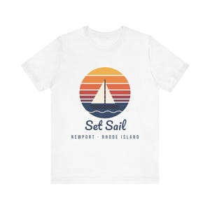 Może przedstawiać: Biały t-shirt z krótkim rękawem z okrągłą grafiką przedstawiającą żaglówkę na wodzie na tle zachodu słońca w pomarańczowe, czerwone i fioletowe paski. Poniżej grafiki znajduje się napis Set Sail oraz NEWPORT · RHODE ISLAND.