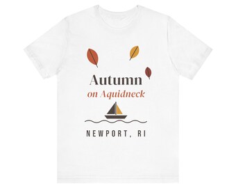 May include: A white short-sleeve t-shirt featuring a graphic with the text Autumn on Aquidneck and NEWPORT, RI. The design includes three falling leaves in orange, yellow, and brown, and a stylised sailboat icon above wavy lines.