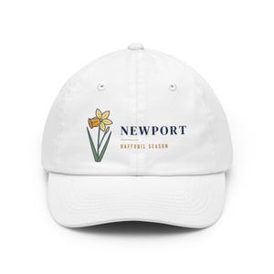 May include: A white baseball cap featuring a graphic of a yellow daffodil with green stems and leaves. To the right of the flower, the text reads NEWPORT in dark blue, with the words DAFFODIL SEASON printed in gold below a horizontal line.