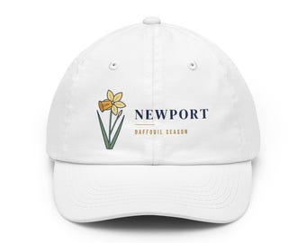 May include: A white baseball cap featuring a graphic of a yellow daffodil with green stems and leaves. To the right of the flower, the text reads NEWPORT in dark blue, with the words DAFFODIL SEASON printed in gold below a horizontal line.