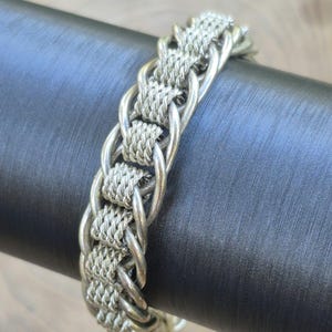May include: A silver-toned metal bracelet featuring a repeating pattern of smooth, interlocking circular links connected by segments of tightly woven, textured wire mesh, creating a continuous, flexible, and detailed chain design.